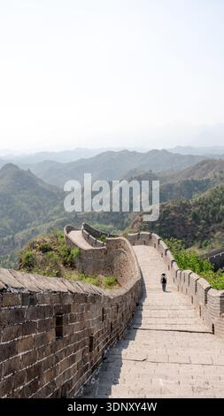 A lone traveler walks along the historic Jinshanling section of the Great Wall in Beijing, surrounded by lush hills and centuries-old brickwork under Stock Photo