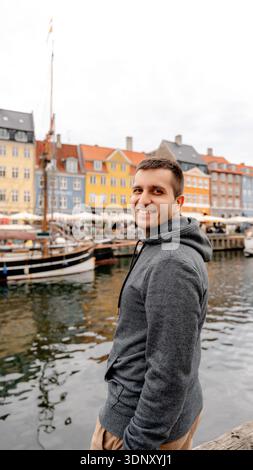 A cheerful man smiles by the picturesque canal in Nyhavn, Copenhagen, Denmark. Vibrant historic buildings and moored boats create a lively, colorful a Stock Photo