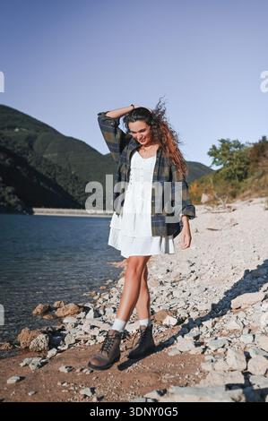 A woman joyfully walks along a picturesque lake in Italy on a sunny day. Wearing a white dress and checkered jacket, she enjoys the tranquility of the Stock Photo