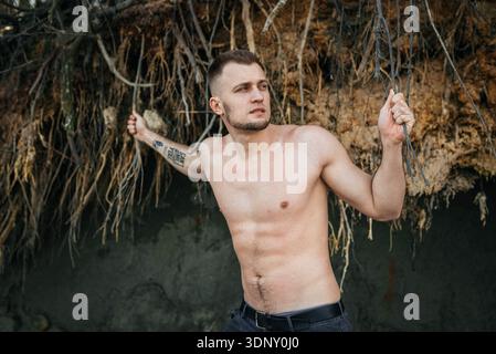 A young man stands confidently against a backdrop of raw nature, with trees and sand. This image captures the untamed beauty and freedom of a secluded Stock Photo