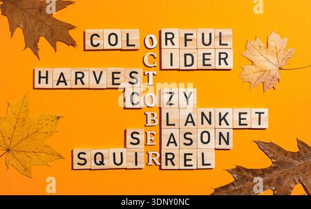 Wooden blocks form a crossword puzzle with autumn words like October and harvest on a vibrant orange background with scattered fall leaves, capturing Stock Photo