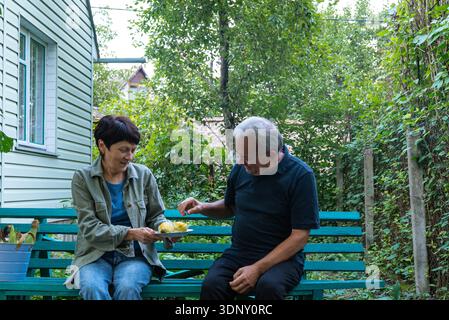 A middle-aged couple sits on a green bench in their lush garden, enjoying freshly harvested corn. The joy of homegrown produce and togetherness is evi Stock Photo