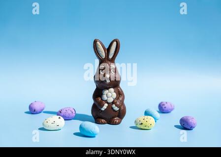 A chocolate Easter bunny with a floral design stands on a light blue background. Vibrant, speckled Easter eggs in pastel colors create a festive, joyf Stock Photo