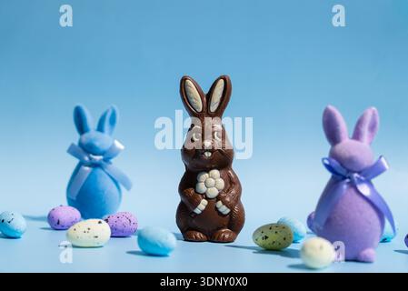 A chocolate Easter bunny stands proudly amidst colorful blue and purple felt bunnies with ribbons. Brightly colored speckled eggs are scattered on a p Stock Photo