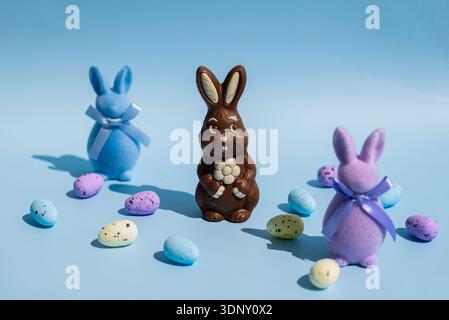 Creative still life featuring a chocolate Easter bunny surrounded by pastel-colored decorative eggs and plush bunnies with blue and purple ribbons on Stock Photo