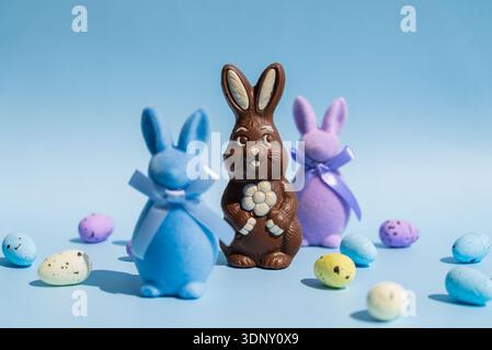 Chocolate Easter bunny with a flower stands surrounded by blue and purple felt bunnies on a pastel blue background. Colorful eggs are scattered around Stock Photo