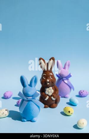 A creative still life of a chocolate Easter bunny surrounded by two pastel-colored bunnies and speckled eggs on a light blue background, evoking festi Stock Photo