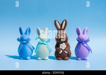 A chocolate Easter bunny with a flower stands amidst three plush pastel bunnies adorned with blue ribbons, set against a bright blue background, showc Stock Photo