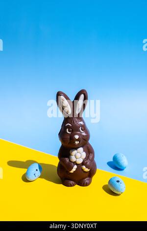 A delightful chocolate Easter bunny with a white flower, set against a vibrant backdrop of blue and yellow. Two blue candy eggs complete this festive Stock Photo