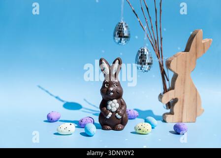 A charming chocolate Easter bunny stands amidst pastel-colored speckled eggs on a light blue background. Wooden bunny and disco ball eggs add a whimsi Stock Photo