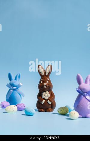 A chocolate Easter bunny stands surrounded by colorful pastel eggs and felt rabbits with bows on a vibrant blue background, creating a festive still l Stock Photo