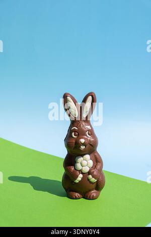 A charming chocolate Easter bunny with a white flower, set against a vibrant green and blue background. The whimsical still life catches the essence o Stock Photo