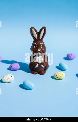 A chocolate Easter bunny holding a flower surrounded by colorful speckled eggs on a bright blue background. The playful arrangement evokes festive and Stock Photo