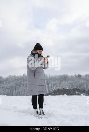 A woman dressed warmly in a gray coat and black hat stands on a snowy landscape, engrossed in her phone. The background features a serene, snow-covere Stock Photo