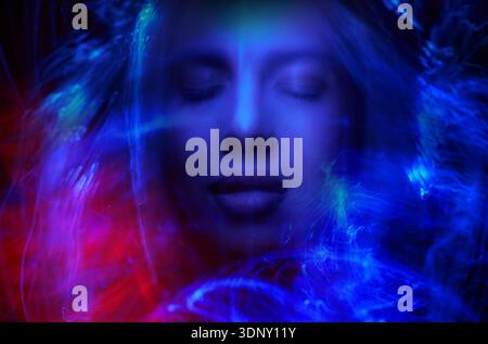 A surreal abstract portrait of a woman with closed eyes, enveloped in ethereal neon blue and red light trails, creating a dreamlike, cosmic atmosphere Stock Photo