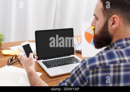 Young beard IT manager looking in mobile phone at work with computer Stock Photo