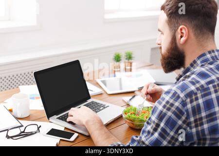 Busy employee eating salad and working with laptop in office, close up Stock Photo