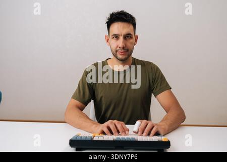 Portrait of young man sitting at white desk and typing on mechanical keyboard, engaged in programming, gaming, general computer use looking at camera Stock Photo
