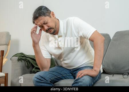 A young man sits on a sofa at home holding his head in pain, representing severe headache, stress, and health problems. Stock Photo