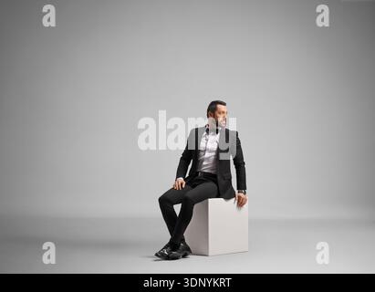 Elegant groom in a black tuxedo sitting on a white cube in a studio Stock Photo