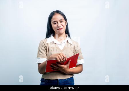 Portrait of a young Southeast Asian woman in a sweater vest holding a red spiral notebook. Smart student or journalist concept in studio shot. Stock Photo