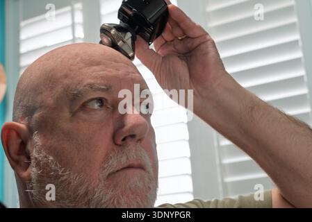 A bald man with a beard is shaving his head with a wire free electric razor. Stock Photo