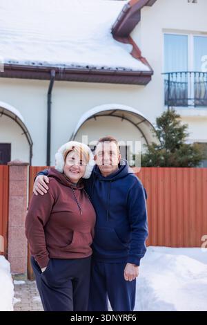 Loving middle aged couple standing together in winter near family home. Happy senior couple hugging outside their house. Healthy aging, lifelong Stock Photo