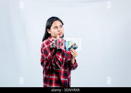 Young Southeast Asian woman in a red flannel shirt looking pensive while holding a fan of colorful credit cards, isolated on a white background. Stock Photo