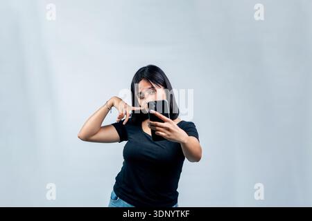 A cheerful young woman in a black t-shirt posing playfully while taking a selfie or recording a video with her smartphone against a white backdrop. Stock Photo