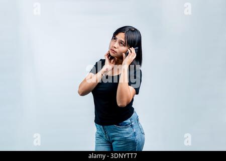 A young woman in a black t-shirt making a phone call, looking up thoughtfully while holding her smartphone against a clean white backdrop. Stock Photo