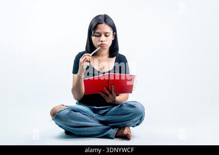 A pensive young woman sitting cross-legged holding a red notebook and putting a pen to her lips, appearing focused or stuck on an idea. Stock Photo