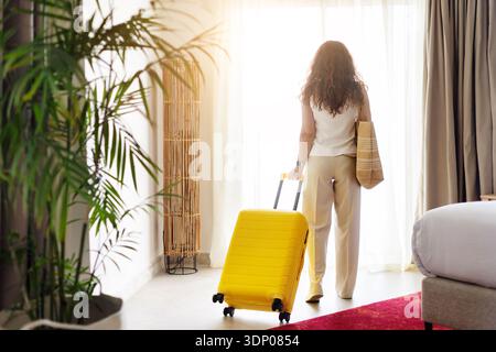 Rear view of woman standing near window with suitcase inside hotel room Stock Photo