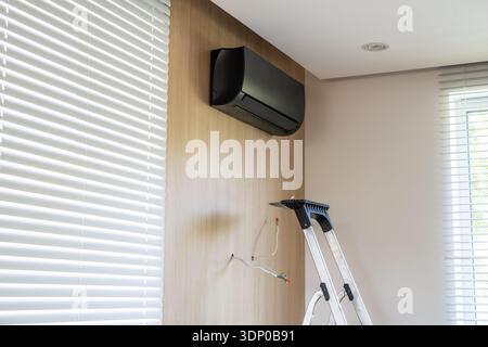 Black air conditioning unit mounted on a wood wall panel in a room with white window blinds and a metal ladder placed beneath it for installation. Stock Photo