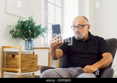 Older man video calling on smartphone, focused expression in bright home Stock Photo