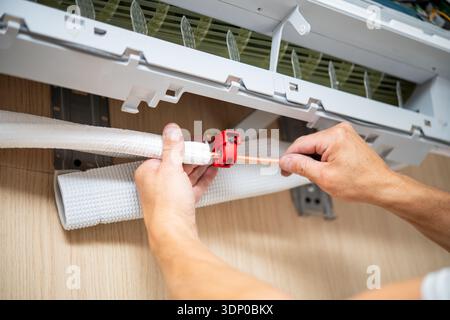 A person installs and secures a copper pipe on a wall-mounted indoor air conditioner with a red clamp in a home setting. Stock Photo