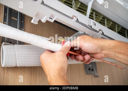 Hands of a technician installing insulation on a copper pipe connected to a domestic wall-mounted air conditioning unit indoors. Stock Photo