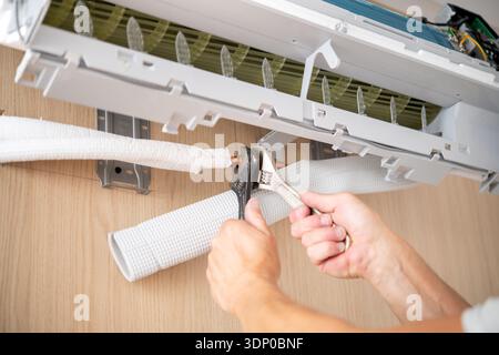 Hands of a technician using pliers and wrench to connect copper pipe to an indoor air conditioner unit mounted on a wooden wall. Stock Photo