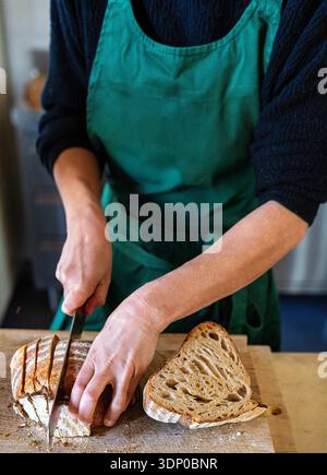 Hands slicing artisan sourdough bread on a wooden cutting board while wearing a green apron in a home kitchen. Stock Photo