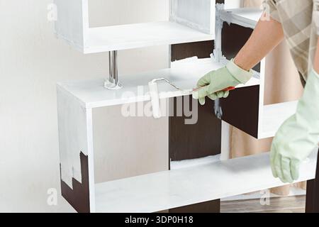Close-up of gloved hand applying white paint to dark wooden furniture shelf using roller, showcasing DIY home renovation, interior design improvement, Stock Photo