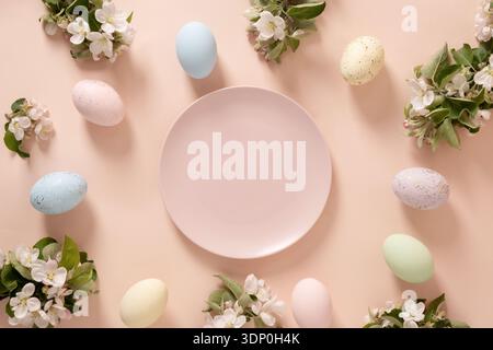 Easter scene with pastel eggs, blooming branches and empty plate in center on soft peach background for festive design. Top view Stock Photo