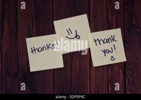 Three Yellow Post-Its with Thank, Thank You and Tongue-Out Smiley on Wooden Table, Top View Flat Lay Stock Photo