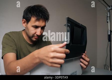 Pensive technician male examining components of disassembled laser printer, troubleshooting and performing maintenance in workshop environment Stock Photo