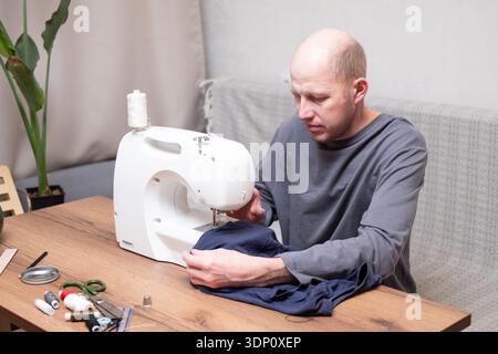 A bald man of 40 years old in a gray T-shirt sits at a table and sews on a sewing machine at home Stock Photo