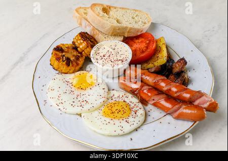 Fried eggs with grilled sausages, corn and white sauce on a plate. A hearty breakfast with two eggs, sausages, tomatoes and slices of bread Stock Photo