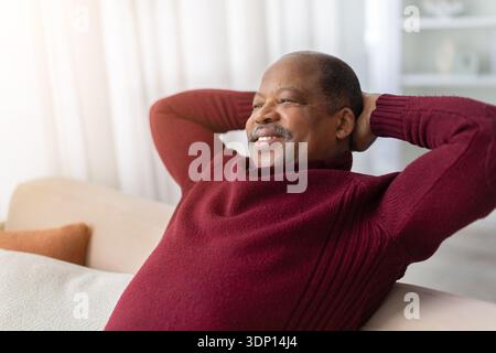 Relaxed black man sitting on sofa at home with hands behind head Stock Photo
