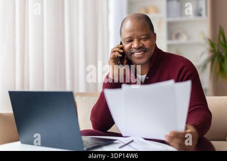 Mature black man talking on smartphone while checking documents at home Stock Photo