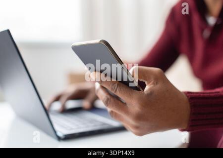 Middle aged black man using smartphone while working on laptop at home Stock Photo