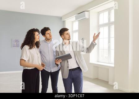 Realtor showing modern house to a young happy couple standing in empty new apartments. Stock Photo