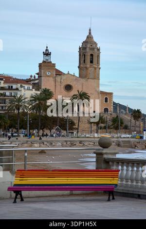 Sitges. Spain - February 12, 2026: Church of Sant Bartomeu and Santa Tecla overlooking the beach, with colorful rainbow bench in foreground on the sea Stock Photo