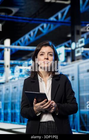 Data center technician using tablet to review equipment diagnostics. IT specialist walking in server hub using digital device to adjust configurations, applying firmware updates, cleaning error states Stock Photo